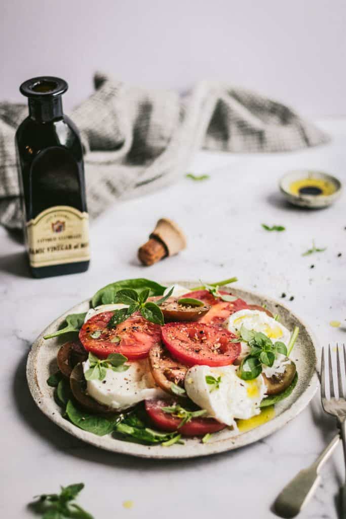 a simple caprese salad on a table with cutlery and a bottle of balsamic vinegar