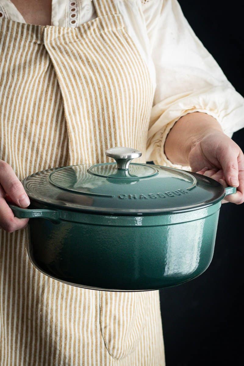 woman in a mustard stipe apron holding a green french oven casserole pot in both hands