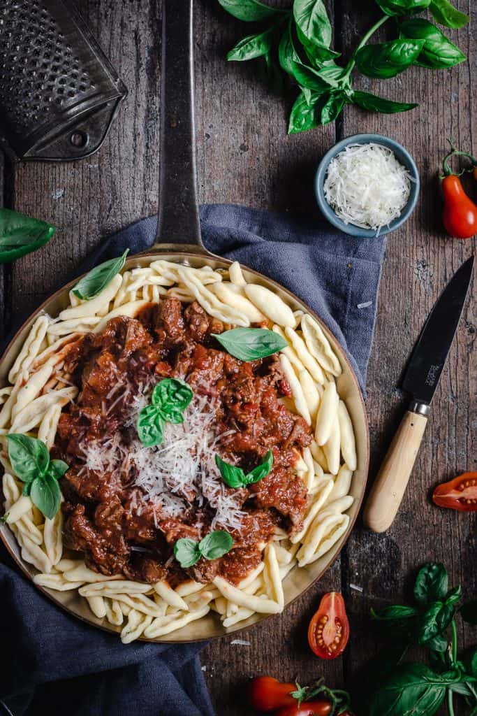flatlay of a frypan of slow cooked beef and red wine ragu on a bed of capunti pasta, with parmasean and basil leaves. 