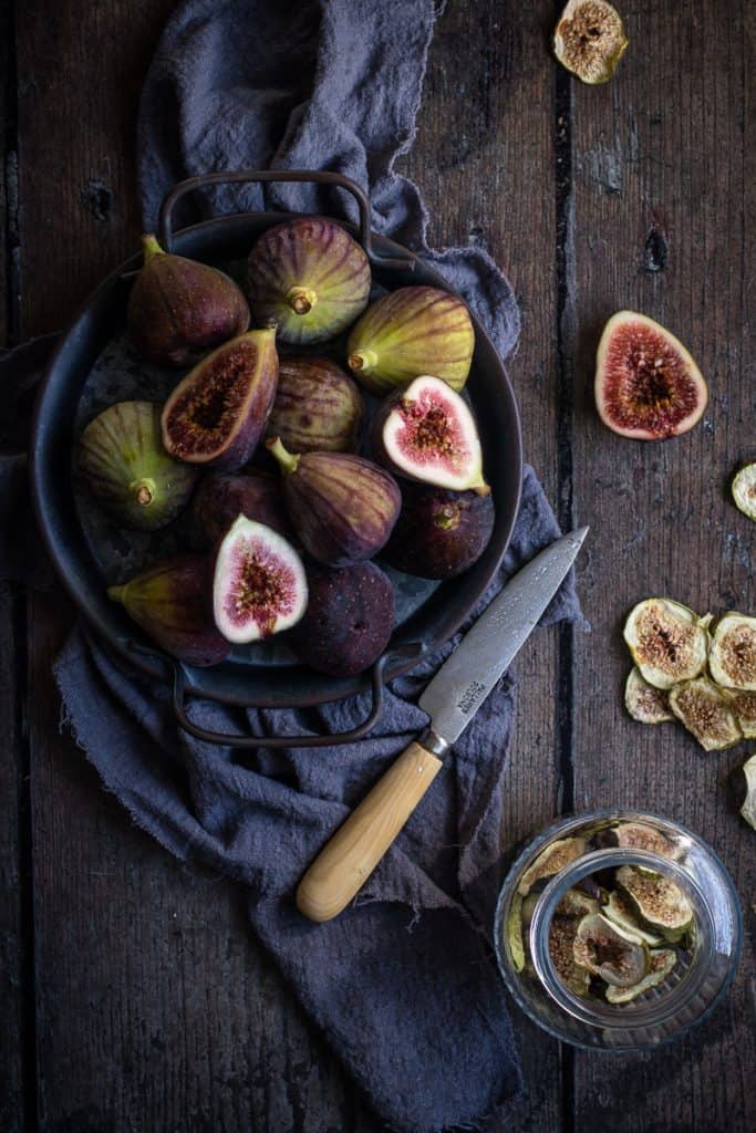 a plate of figs getting cut in half ready for dehydrating. In the bottom corn is a jar of dehydrated figs with a few scattered around