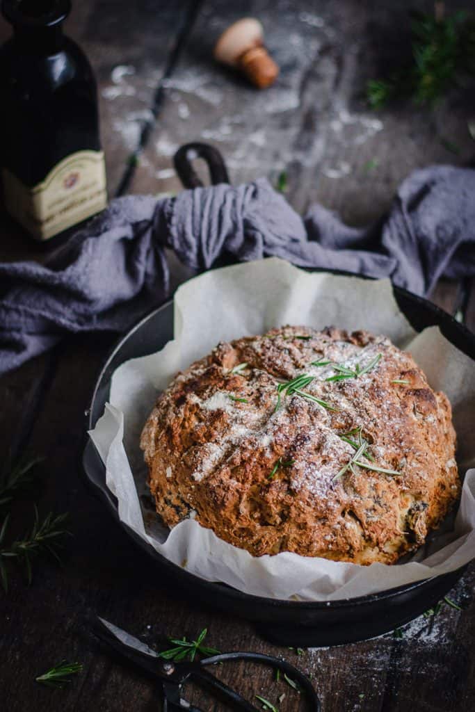 a loaf of rosemary fig and walnut soda bread in a seasoned cast iron skillet on a bench surrounded by scattered rosemary leaves and ingredients.