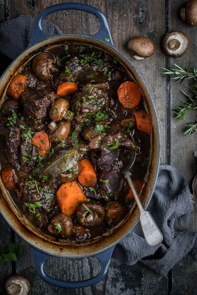 an oval cast iron french oven full of slow cooked beef cheeks in red wine with carrots and mushrooms. 
