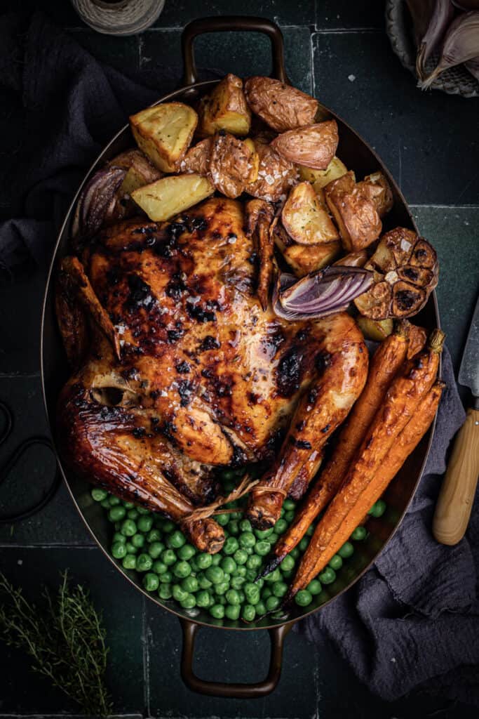 an oval roasting pan with a black garlic roast chicken, roast vegetables and peas in it.