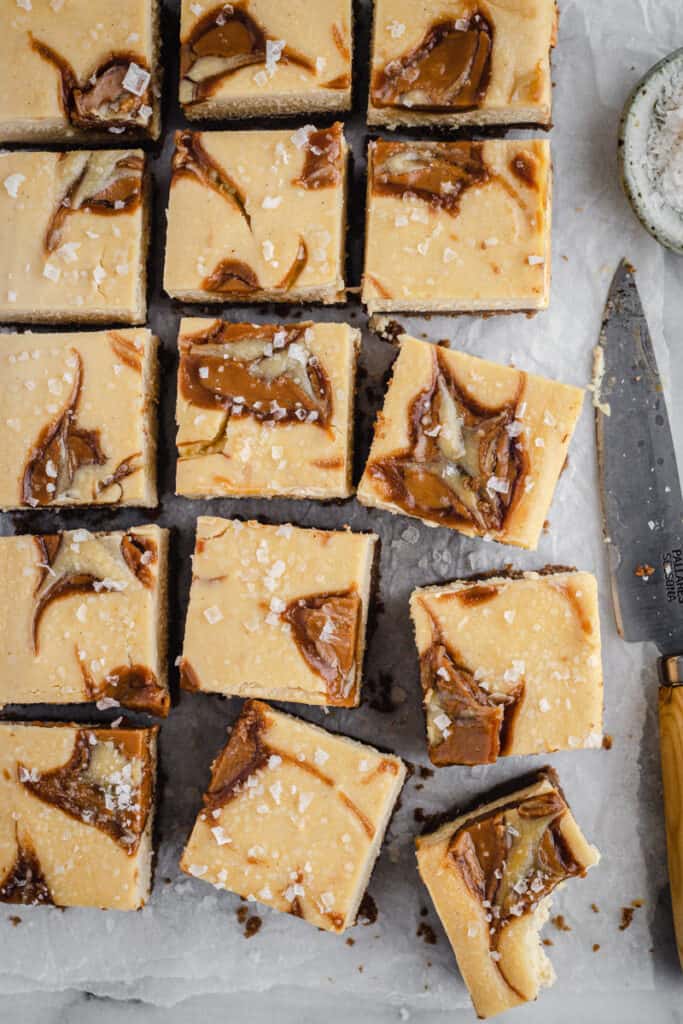 square slices of baked biscoff cheesecake slice on a marble board lined with baking paper. A knife and bowl of sea salt sit beside.