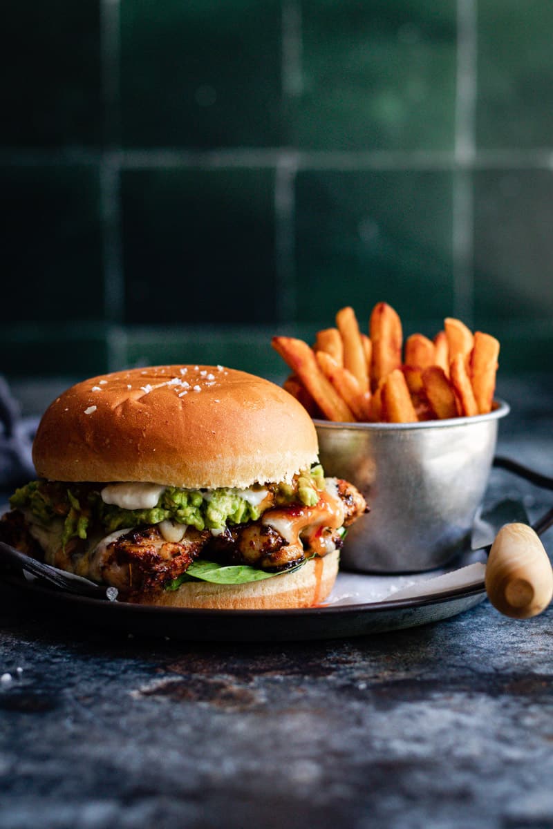 a juicy grilled cajun chicken burger with smashed avocado and sweet chilli sauce alongside a side of fries on a metal tray.