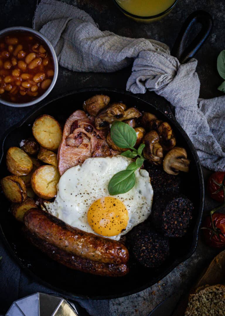a traditional full irish breakfast just cooked in a black cast iron frypan. On a bench alongside baked beans, orange juice and a pot of coffee