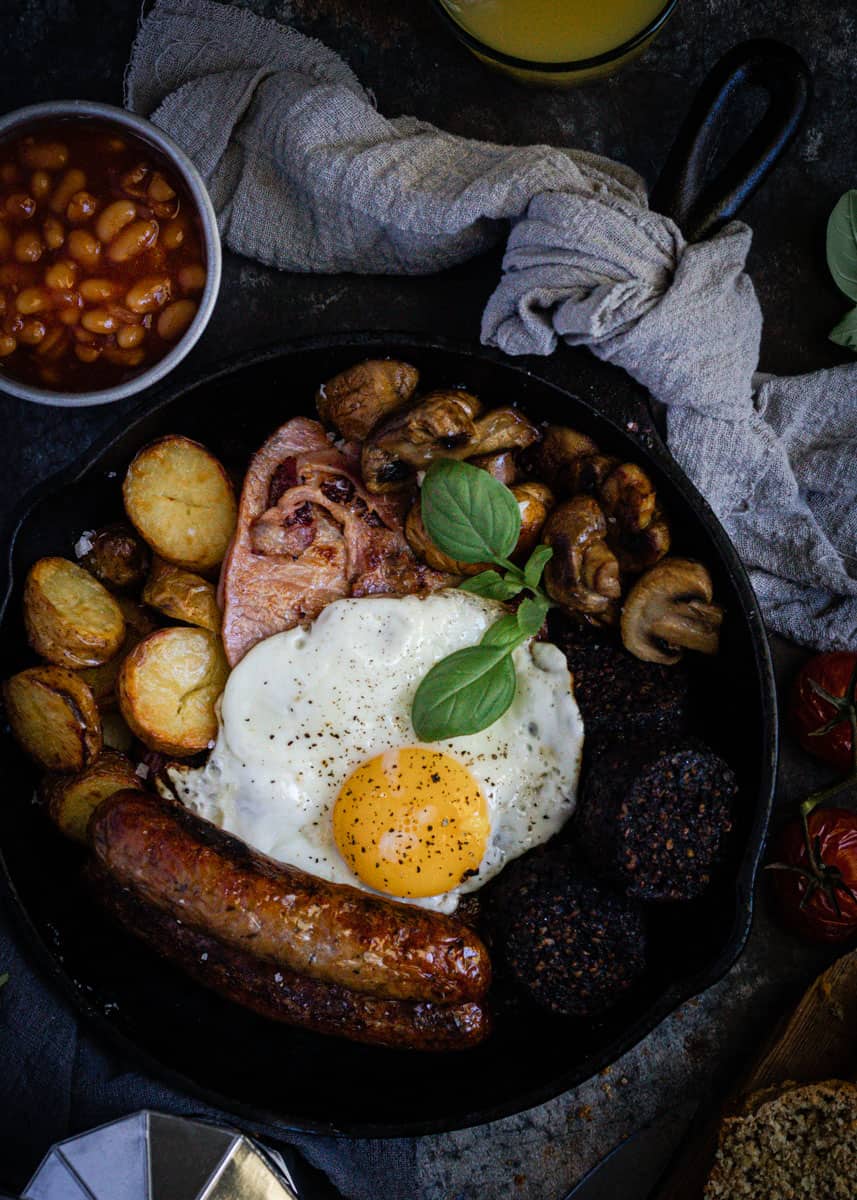 a traditional full irish breakfast just cooked in a black cast iron frypan. On a bench alongside baked beans, orange juice and a pot of coffee