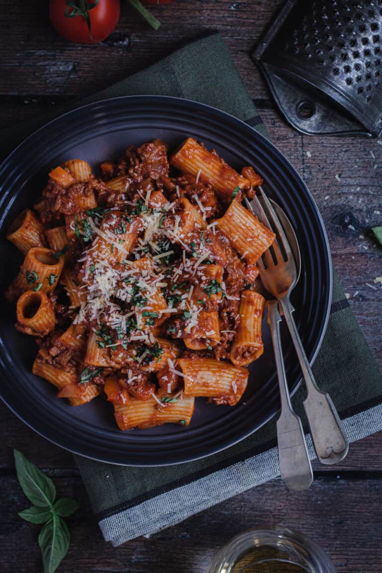 a bowl of rigatoni bolognese sprinkled with parmesan cheese and fresh basil on a wooden table.
