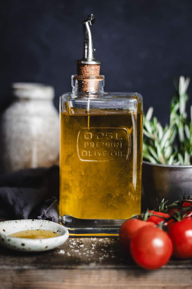 A glass bottle of oil alongside fresh herbs, salt and cherry tomatoes on the vine.