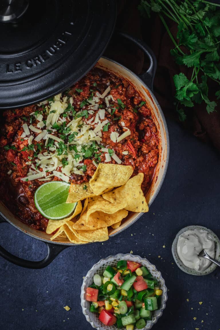 A black dutch oven full of black bean chili con carne with a chopped salad and corn chips.