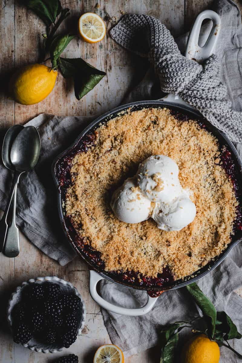 An off white skillet filled with oven baked apple and blackberry crumble topped with three scoops of vanilla ice cream.