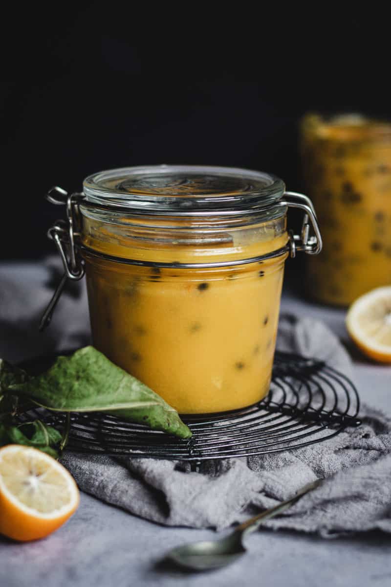 A jar of lemon and passionfruit curd on a wire cooling rack surrounded by small lemons and more curd.