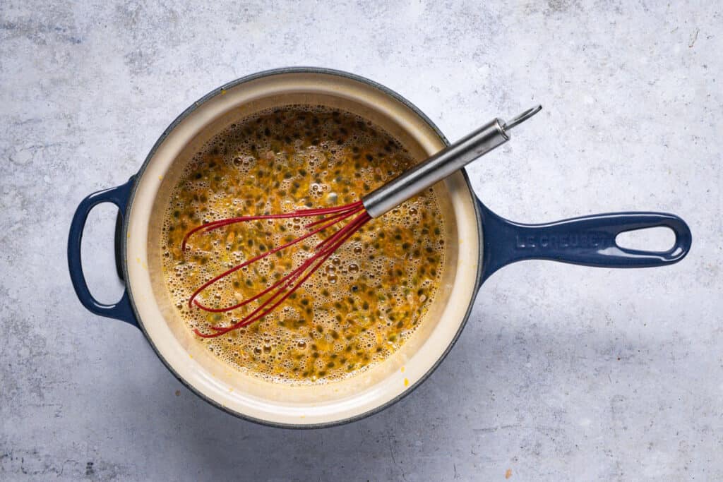 a blue saucepan with a red whisk in it stirring all the lemon and passionfruit curd ingredients together.