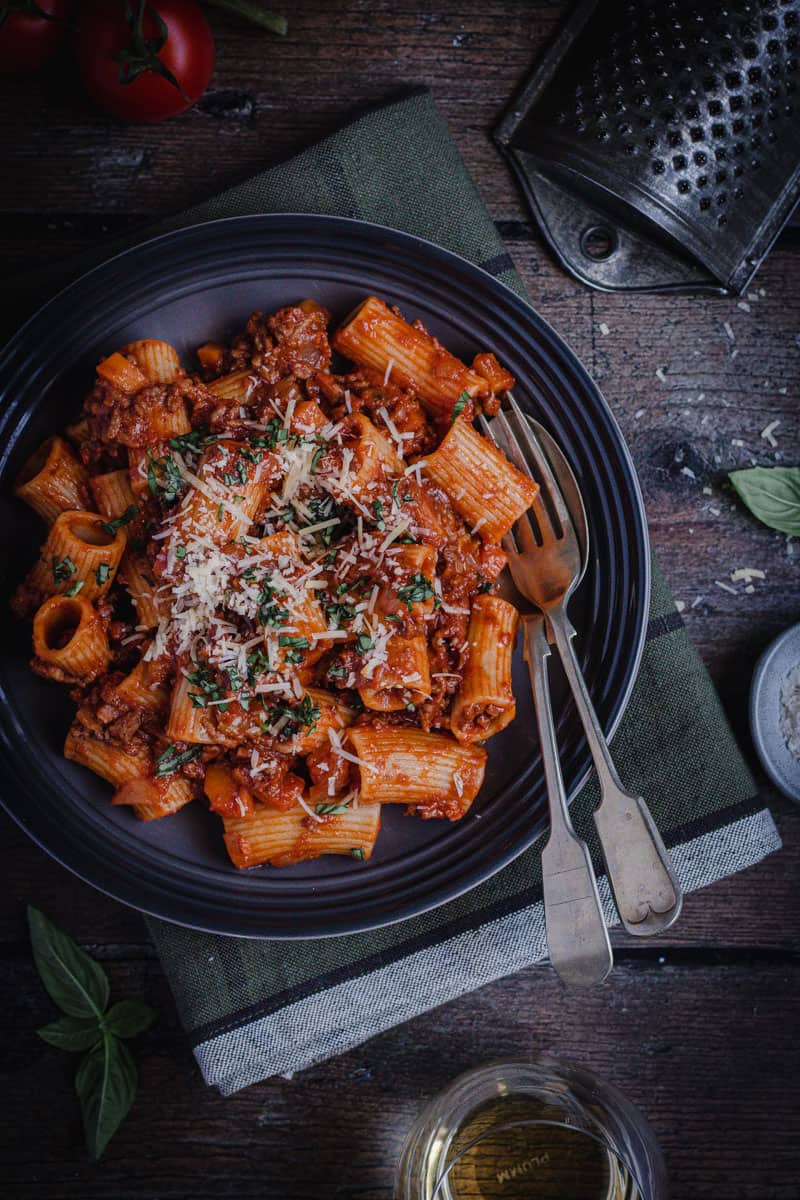 A bowl of slow cooked rigatoni bolognese on a table alongside a glass of wine.