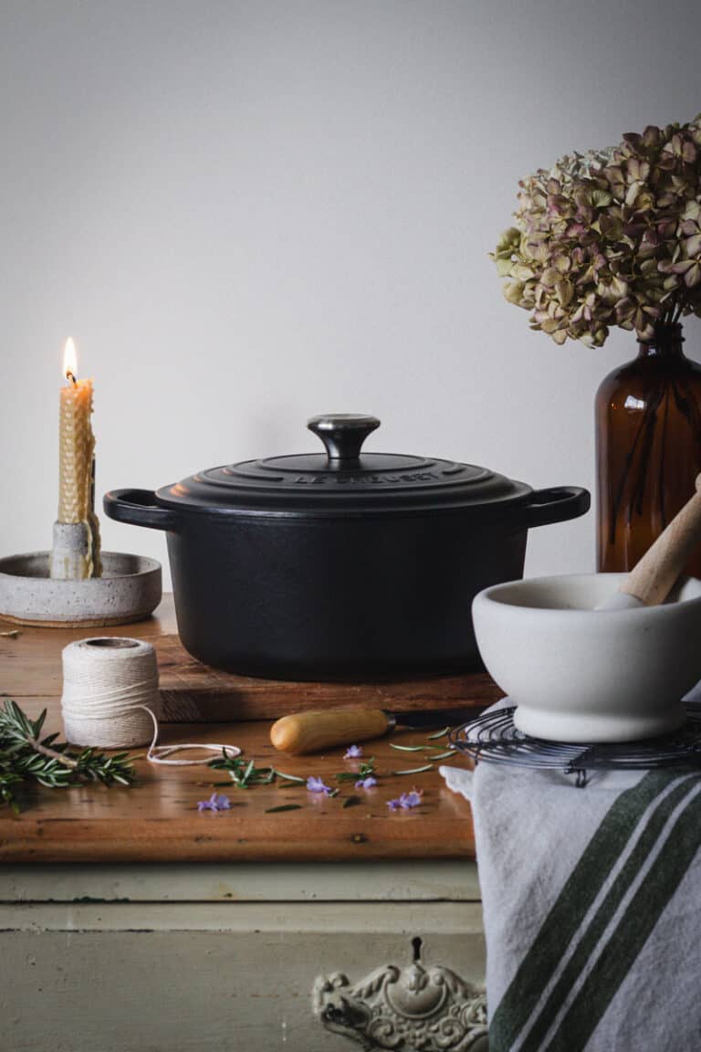 A black le creuset dutch oven sitting on a kitchen table surrounded by cooking bits and pieces.