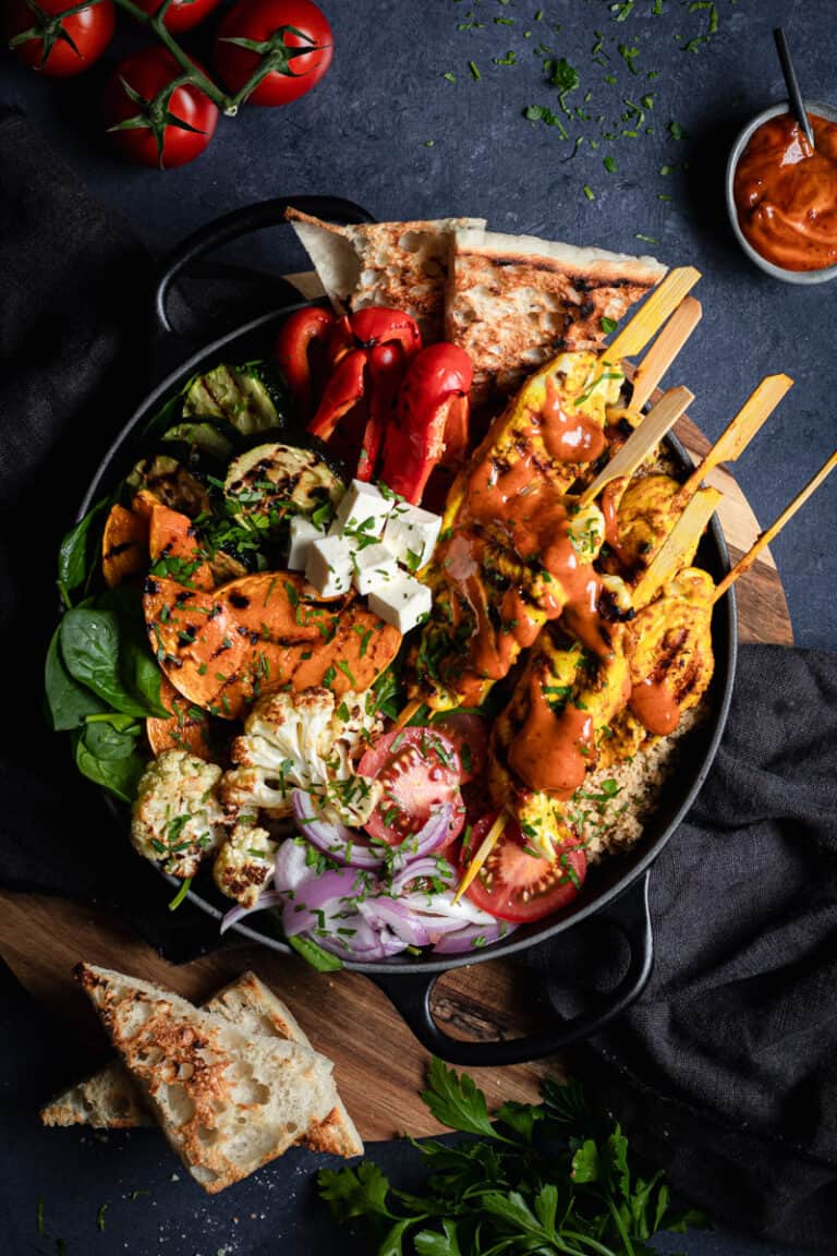 A Black cast iron shallow casserole, with roasted vegetables and curried chicken kebab salad. There is some toasted Turkish bread on the side.