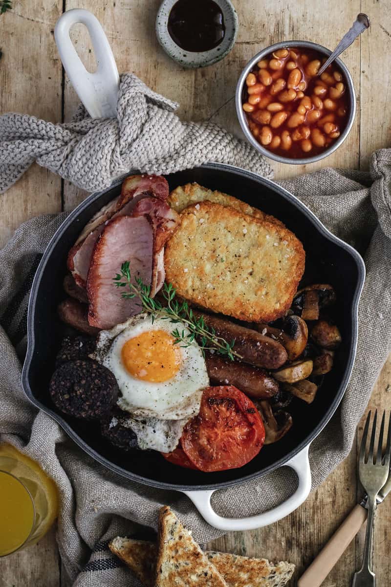 A traditional full irish breakfast just cooked in a cast iron frypan. On a bench alongside baked beans, orange juice and brown sauce.