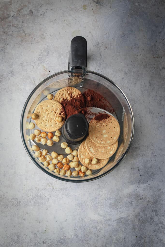 Roasted hazelnuts, Marie biscuits and dutch cocoa in a food processor bowl ready to chop.
