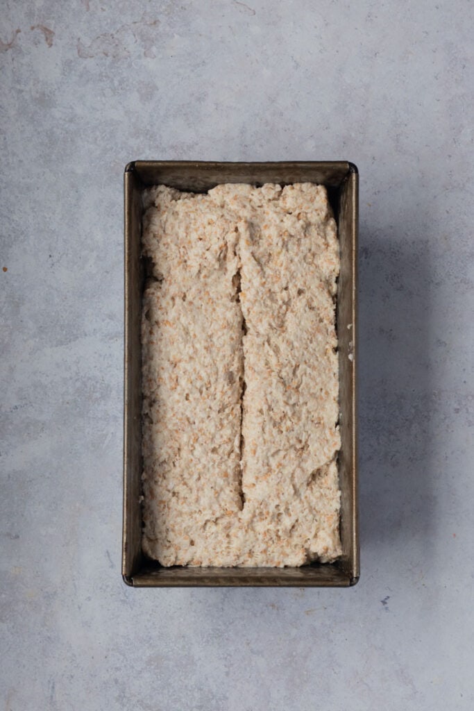 The dough is in a metal loaf tin. The center of the loaf has been scored with a vertical line.