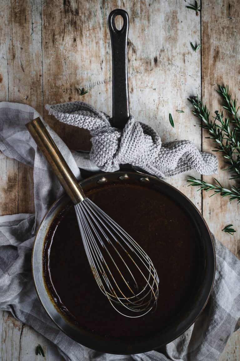 A copper frying pan full of roast lamb gravy. A whisk sits inside the frying pan and fresh rosemary if beside the pan.