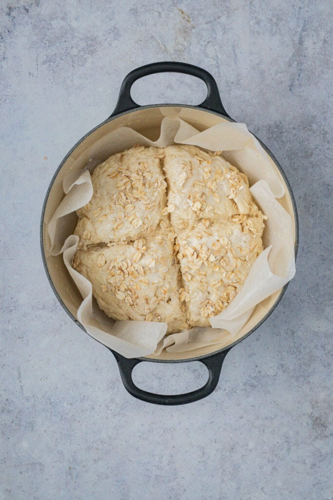 The load of soda bread is in a black cast iron Dutch oven. It is scored and dusted with oats.